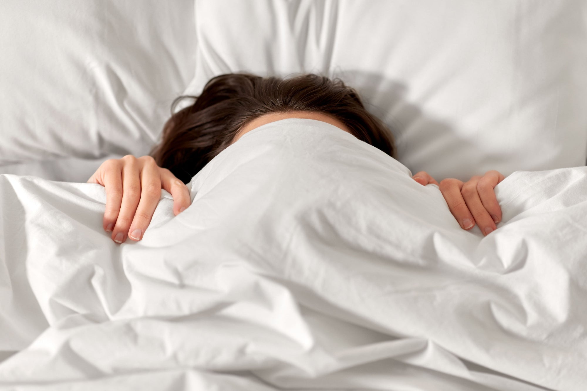 Femme sous une couette blanche qui se cache le visage avec parce qu'elle ne veut pas se lever et elle est fatiguée