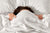 Femme sous une couette blanche qui se cache le visage avec parce qu'elle ne veut pas se lever et elle est fatiguée
